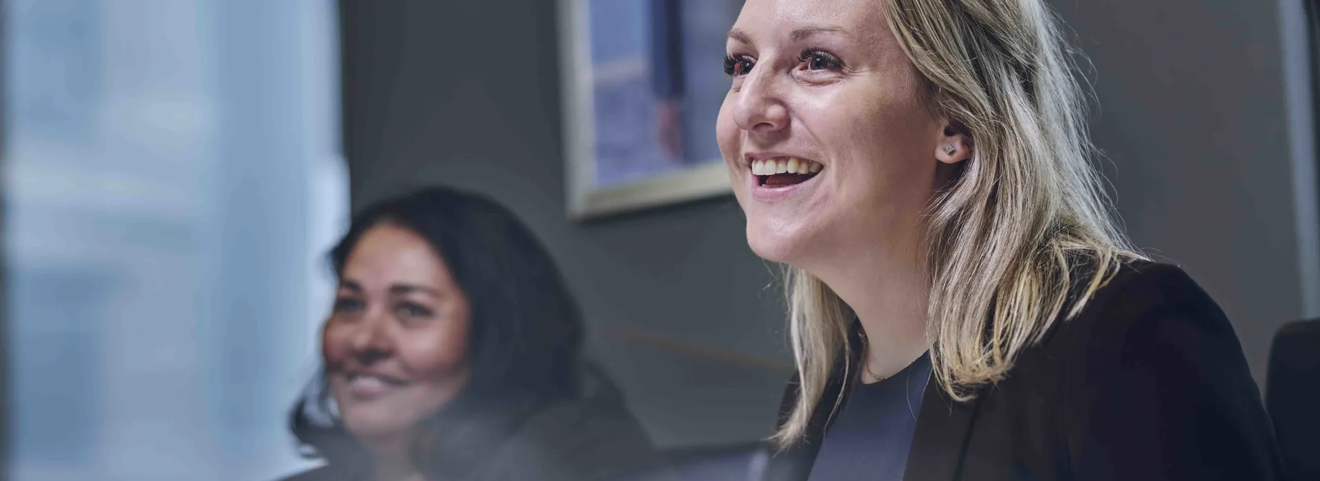 Two women seated together in a professional setting, one in the foreground smiling widely.