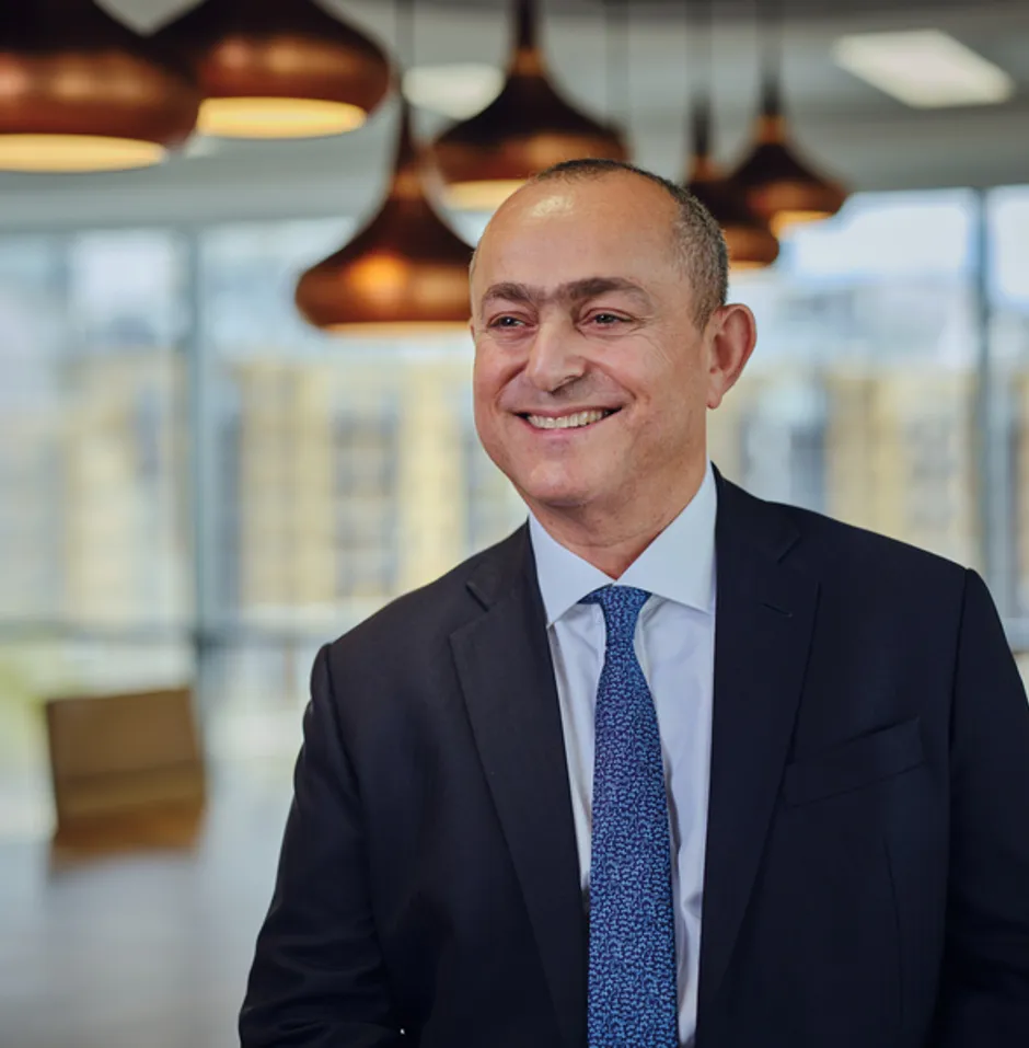 Smiling man in a dark suit standing indoors with a cityscape view through large windows.