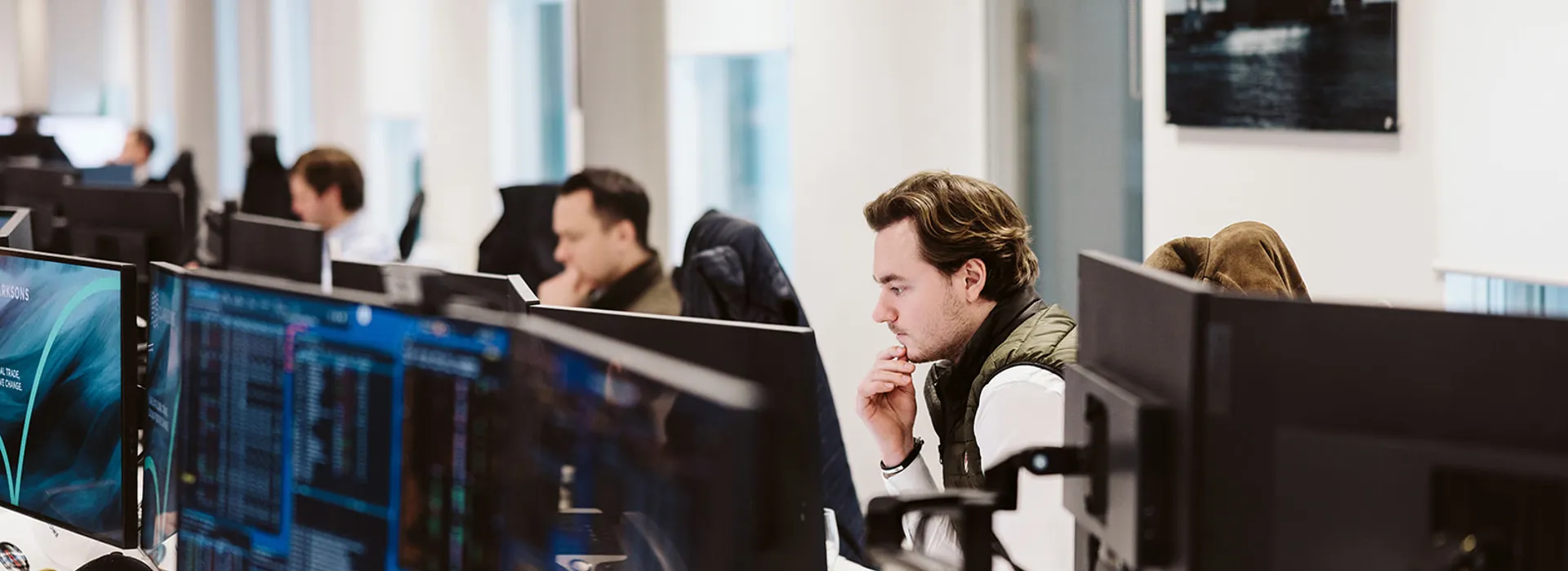 Modern office space with employees focused on computer screens; a man in a green vest rests his chin on his hand.