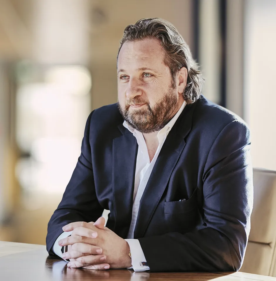 Man with a beard in a dark suit seated at a table, deep in thought, in an office setting.