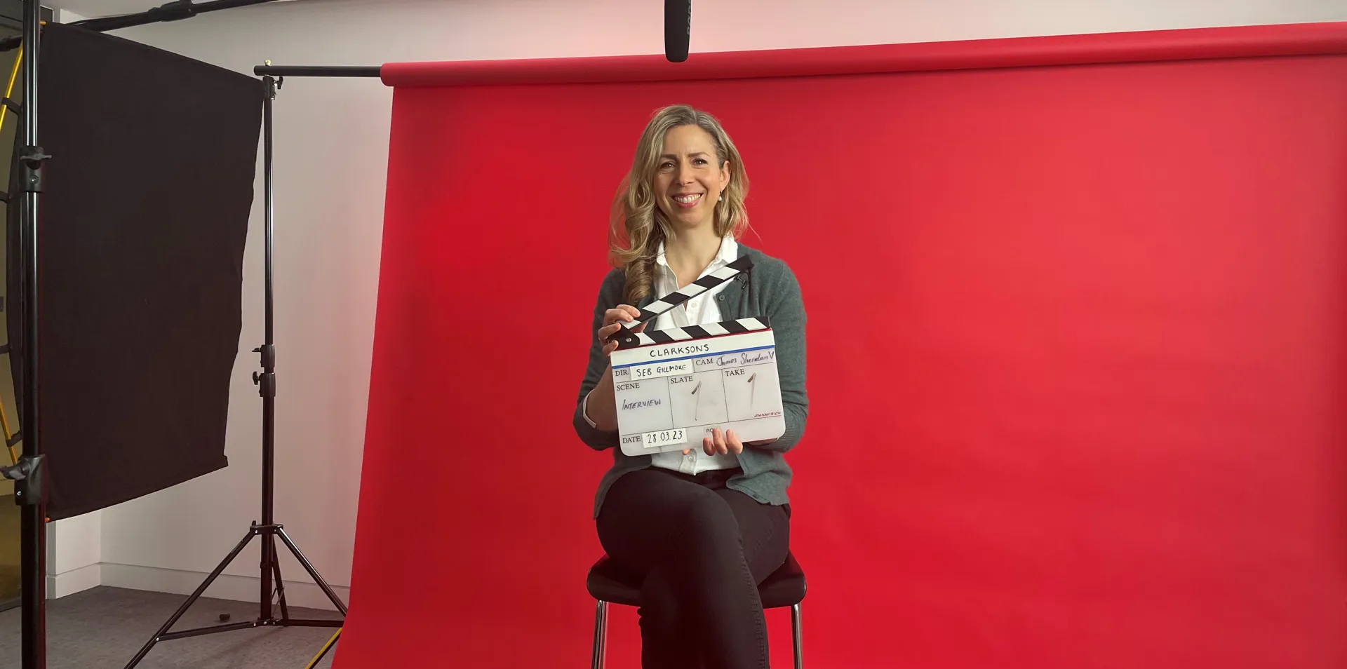 woman sitting on a stool behind a red background, holding a clapper