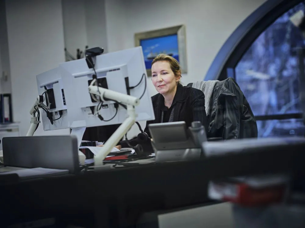 Woman in a black jacket seated at a modern desk focused on two computer monitors.