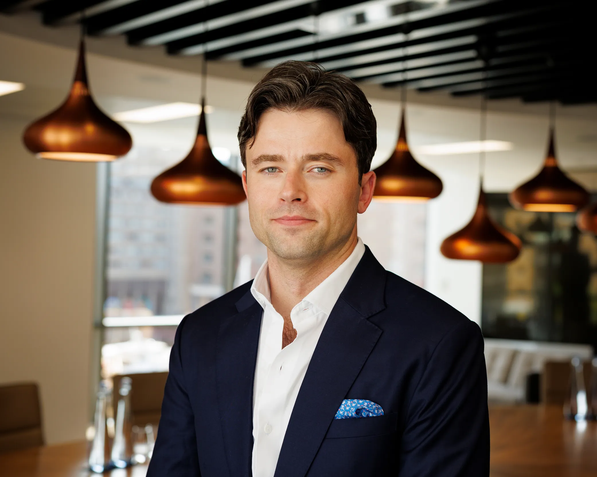 Man in a navy suit jacket with a pocket square standing indoors with an urban background.