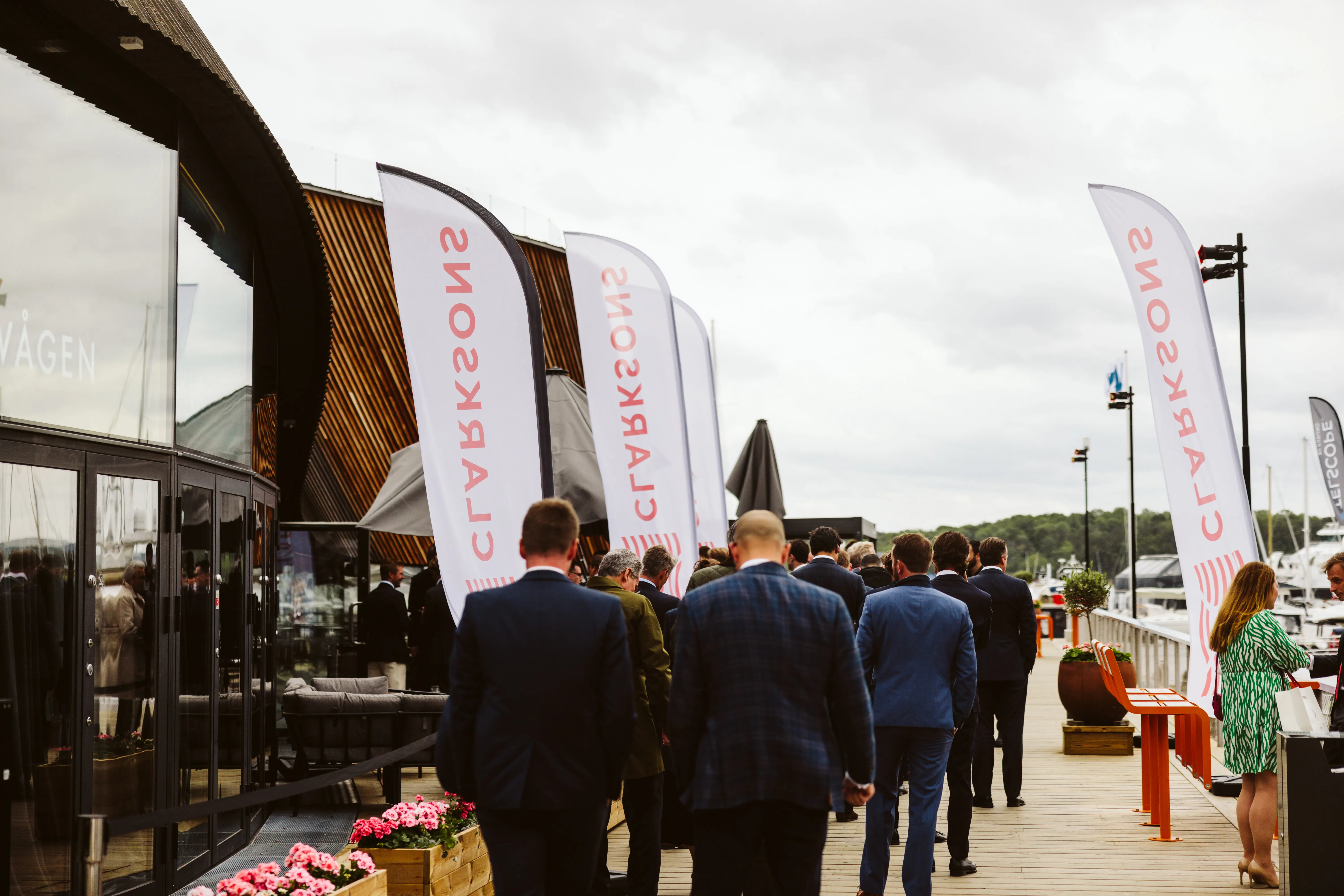 Group in formal attire walking on a deck beside a marina with DEOLS flags.