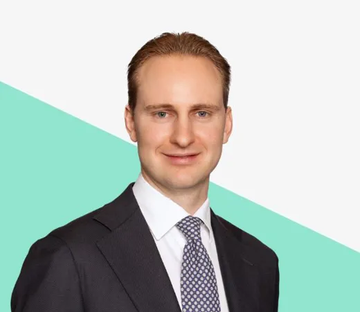 Smiling man in a dark suit and tie against a light background with a mint green diagonal shape.
