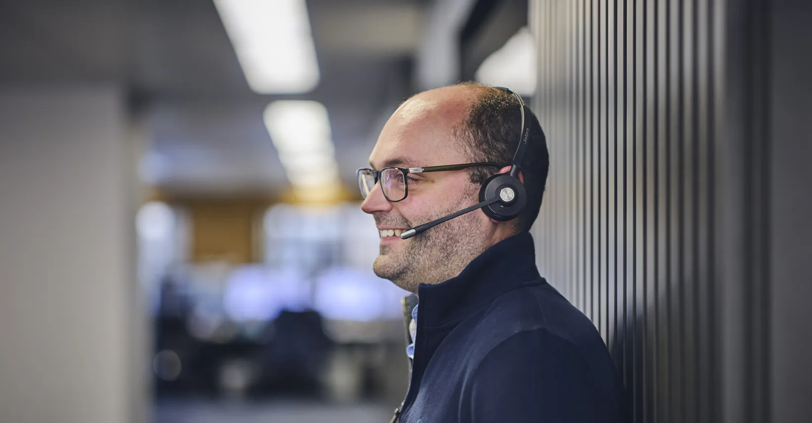 Smiling man with glasses and a headset in an office environment.