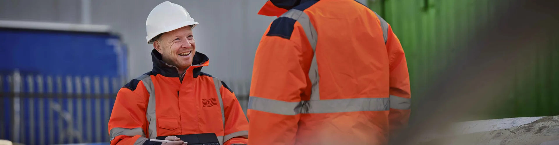 Two workers in orange jackets discussing outdoors.
