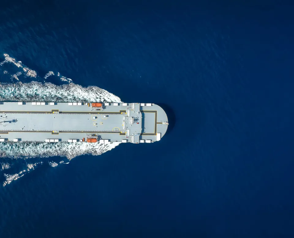 Aerial view of a large cargo ship moving through deep blue water, leaving a white wake.