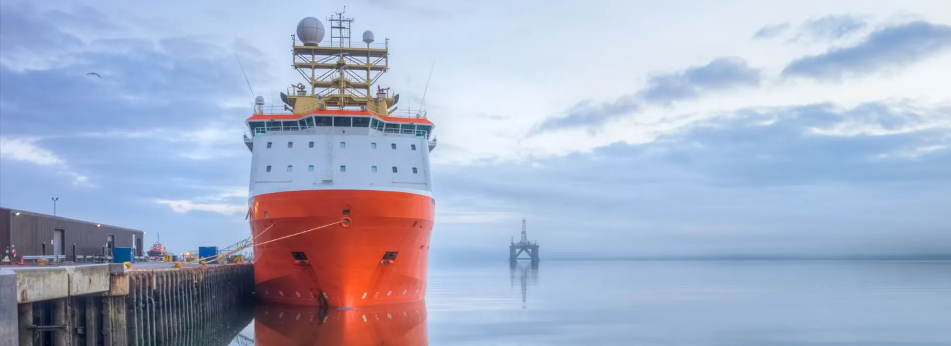 Large white and orange ship docked at a pier, reflecting in calm water, with an oil rig on the horizon.