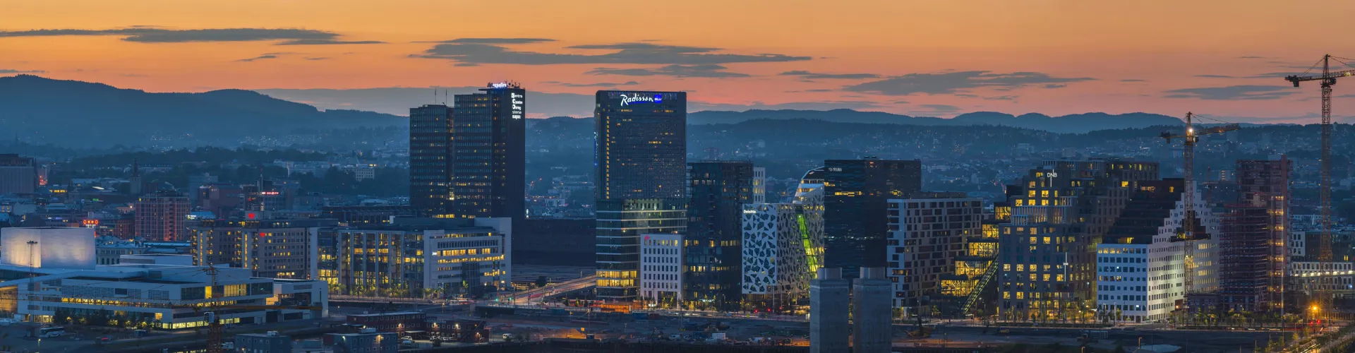 Panoramic view of modern city skyline at dusk with illuminated high-rise buildings and distant mountains.