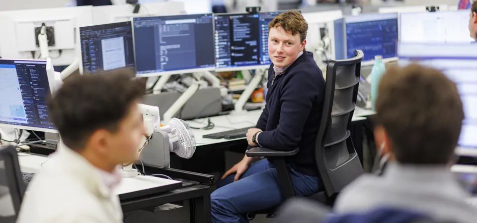 Young man in a dark sweater in an office chair looking at colleagues with monitors showing code/data.