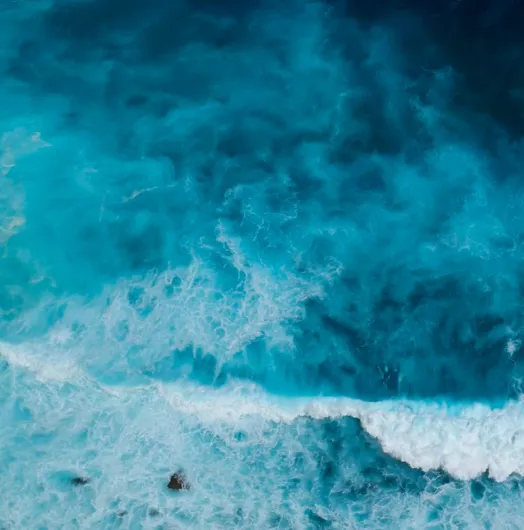 Aerial view of turquoise ocean with white waves crashing near rocky shore.
