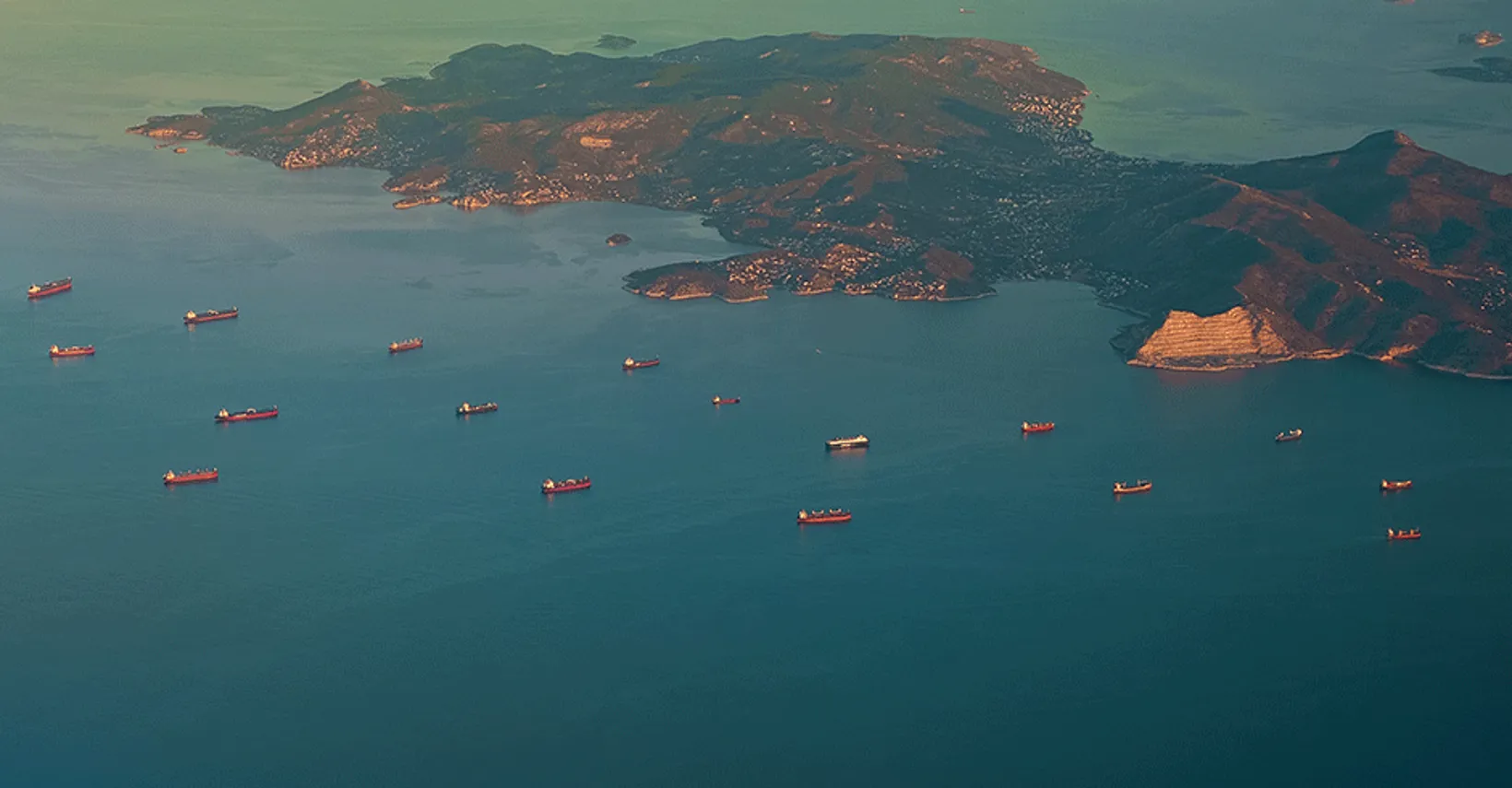 Aerial view of a coastal area with cargo ships, a mountainous island, and a tranquil blue sea.