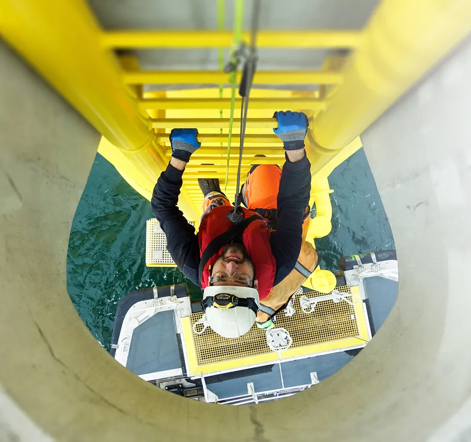 Manual high worker offshore climbing on wind-turbine on ladder and transfer vessel waiting under wind turbine