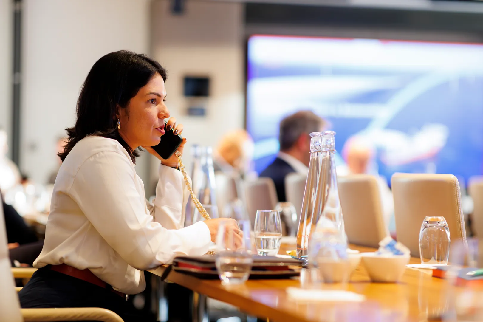 Woman in a white blouse sitting at a conference table talking on a phone.