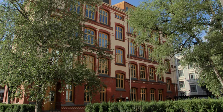 Multi-story red-brick building with large windows, decorative details, and surrounding trees.