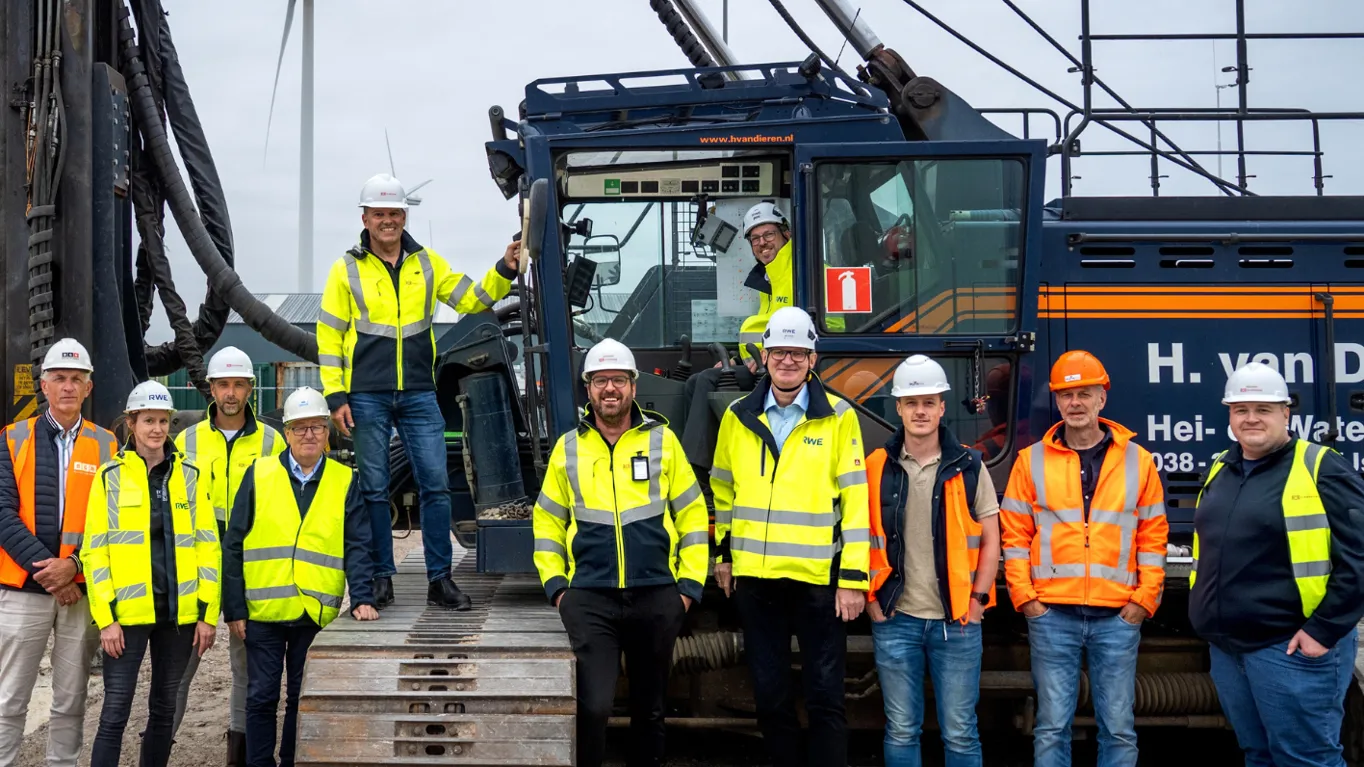 Group of 11 in safety gear posing by heavy machinery with a wind turbine backdrop.
